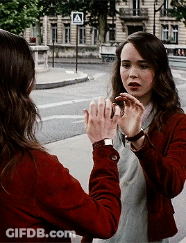 A Woman In A Red Jacket Talks To Another Woman In Front Of A Crosswalk Sign That Says A GIF