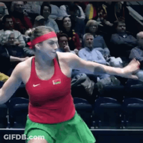 A Woman In A Red Nike Tank Top And Green Skirt Is Playing Tennis In Front Of A Crowd GIF