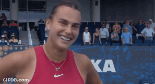 A Woman In A Red Nike Tank Top Smiles In Front Of A Ka Sign GIF