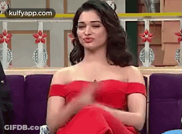 A Woman In A Red Off The Shoulder Dress Is Sitting On A Couch Making A Funny Face . GIF