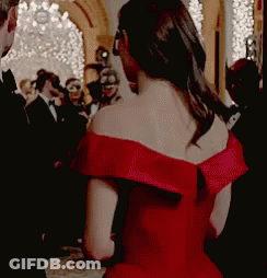 A Woman In A Red Off The Shoulder Dress Is Standing In Front Of A Crowd . GIF