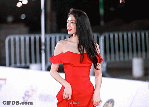 A Woman In A Red Off The Shoulder Dress Stands On A White Carpet GIF