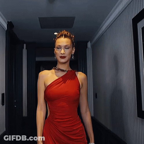 A Woman In A Red One Shoulder Dress Walks Down A Hallway GIF