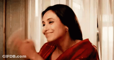 A Woman In A Red Saree Is Smiling And Clapping Her Hands In Front Of A Window . GIF