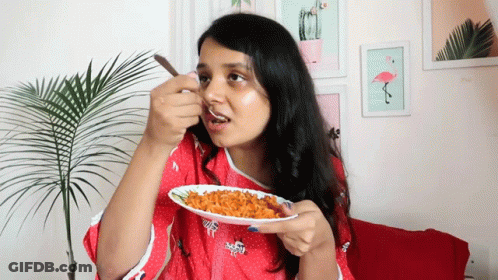 A Woman In A Red Shirt Is Eating A Plate Of Food With A Spoon GIF