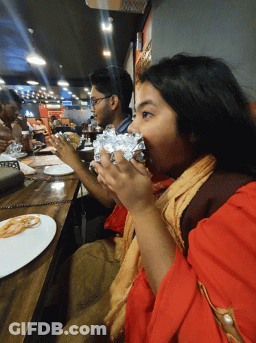 A Woman In A Red Shirt Is Eating A Wrapped Sandwich In A Restaurant GIF
