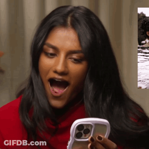 A Woman In A Red Shirt Is Holding A Cell Phone With Her Mouth Open GIF