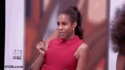 A Woman In A Red Shirt Is Sitting In Front Of A Wall And Making A Funny Face . GIF