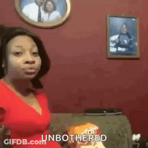 A Woman In A Red Shirt Is Sitting On A Couch Holding A Bag Of Chips And Making A Funny Face . GIF