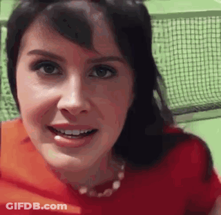 A Woman In A Red Shirt Is Smiling And Looking At The Camera While Standing In Front Of A Tennis Net . GIF