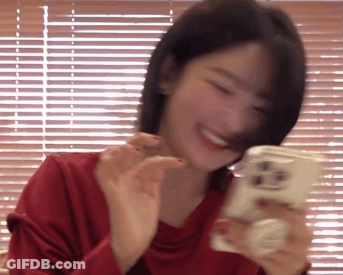 A Woman In A Red Shirt Is Smiling While Holding A Cell Phone In Front Of A Window With Blinds . GIF