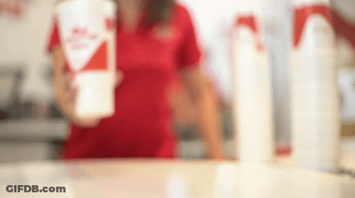 A Woman In A Red Shirt Is Standing Behind A Counter Holding A Cup Of Milkshake . GIF