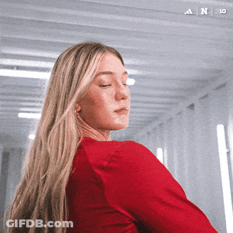 A Woman In A Red Shirt Is Standing In A Hallway With The Adidas Logo On The Bottom Left GIF