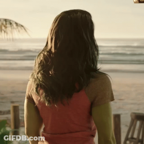 A Woman In A Red Shirt Is Standing On A Beach Looking At The Ocean GIF