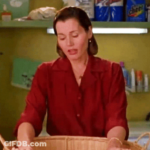 A Woman In A Red Shirt Is Washing Clothes In A Laundry Room With A Bag Of Cheer On The Shelf Behind Her GIF