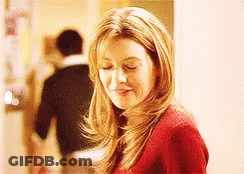 A Woman In A Red Sweater Is Smiling With Her Eyes Closed And A Man In A Black Shirt Is Walking Behind Her . GIF