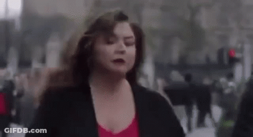 A Woman In A Red Top And Black Jacket Is Running Down A City Street . GIF
