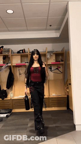 A Woman In A Red Top And Black Pants Is Standing In A Locker Room Holding A Water Bottle GIF