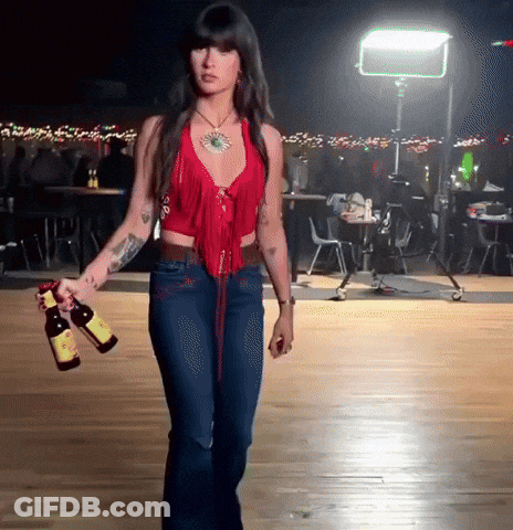 A Woman In A Red Top And Blue Jeans Holding Two Bottles Of Beer GIF