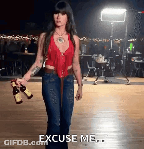 A Woman In A Red Top And Blue Jeans Is Holding Two Bottles Of Wine And Says Excuse Me GIF