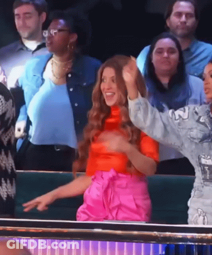 A Woman In A Red Top And Pink Pants Stands In A Crowd Of People GIF