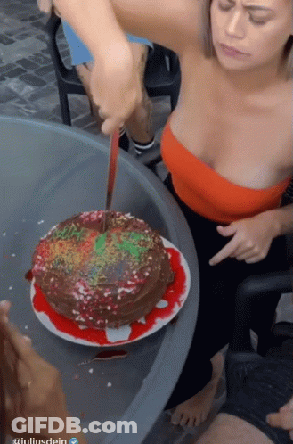 A Woman In A Red Top Is Cutting A Cake With A Knife In Front Of A Sign That Says Giuliardein GIF