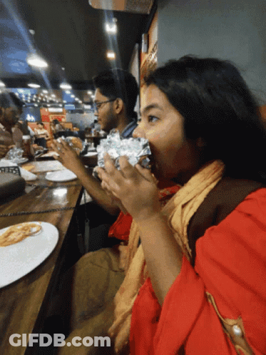 A Woman In A Red Top Is Eating A Wrapped Sandwich GIF