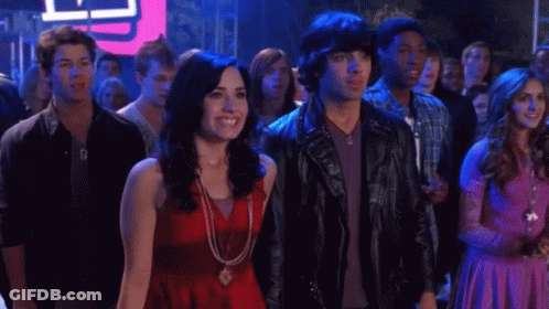 A Woman In A Red Top Stands Next To A Man In A Leather Jacket With Studs On It GIF