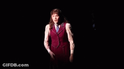 A Woman In A Red Vest And White Shirt Is Standing On A Stage In The Dark GIF