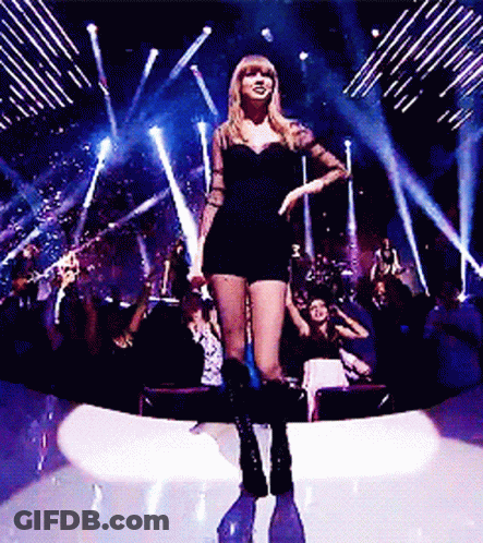 A Woman In A Short Black Dress Is Standing On A Stage In Front Of A Crowd GIF