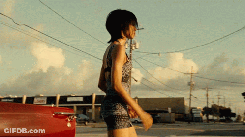 A Woman In A Short Dress Is Walking Down A Street In Front Of A Building That Says ' 365 ' On It GIF