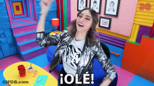 A Woman In A Silver Jacket Is Sitting In A Colorful Room With Her Arms In The Air And Says " Ole " GIF