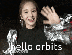 A Woman In A Silver Jacket Is Waving Her Hand And Says Hello Orbits GIF
