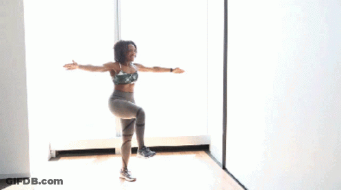 A Woman In A Sports Bra And Leggings Is Standing On One Leg In Front Of A Window With Her Arms In The Air GIF