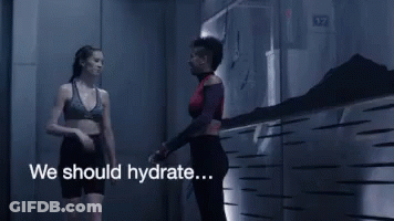 A Woman In A Sports Bra Is Walking Down A Hallway With The Words We Should Hydrate GIF