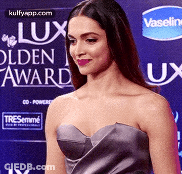 A Woman In A Strapless Dress Is Standing On A Red Carpet In Front Of A Vaseline Sign . GIF