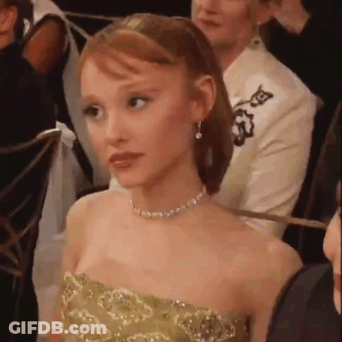 A Woman In A Strapless Gold Dress Is Sitting In A Crowd Of People . GIF