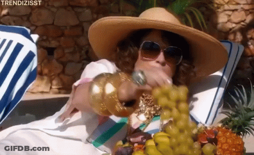 A Woman In A Straw Hat And Sunglasses Is Eating Grapes While Laying On A Beach Towel . GIF