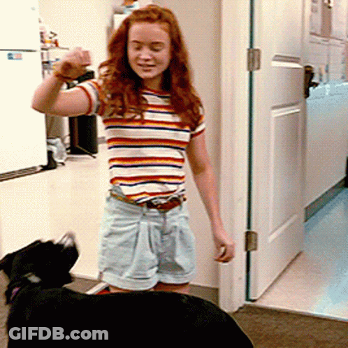 A Woman In A Striped Shirt And Shorts Stands Next To A Black Dog GIF