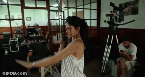 A Woman In A Striped Shirt Is Dancing In A Room With A Camera On A Tripod . GIF