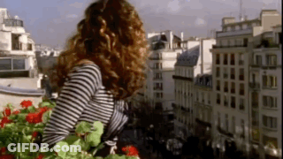 A Woman In A Striped Shirt Is Standing On A Balcony Looking Out Over A City GIF