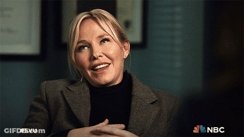 A Woman In A Suit And Black Turtleneck Is Smiling With The Nbc Logo Behind Her GIF