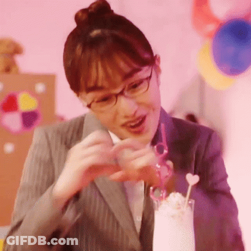 A Woman In A Suit And Glasses Is Making A Heart With Her Hands Next To A Glass Of Milkshake . GIF