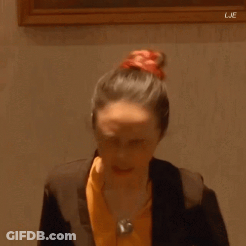 A Woman In A Suit And Orange Shirt Is Standing In Front Of A Framed Painting . GIF