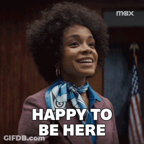 A Woman In A Suit And Scarf Is Smiling And Says " Happy To Be Here " GIF