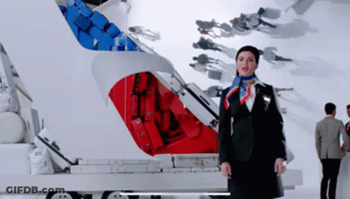 A Woman In A Suit And Scarf Stands In Front Of A Red And Blue Suitcase GIF