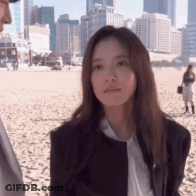 A Woman In A Suit And Tie Is Standing On A Sandy Beach GIF