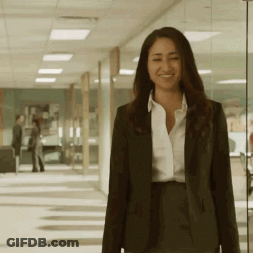 A Woman In A Suit And White Shirt Smiles While Walking Down A Hallway GIF