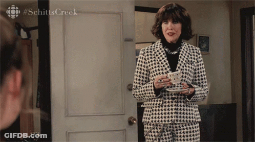 A Woman In A Suit Holds A Cup Of Coffee In Front Of A Door With #Schitts Creek Written On It GIF