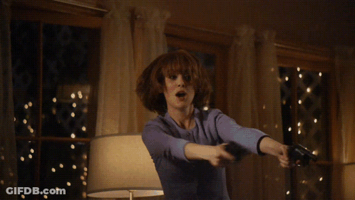 A Woman In A Suit Is Holding Two Guns In Front Of A Table GIF